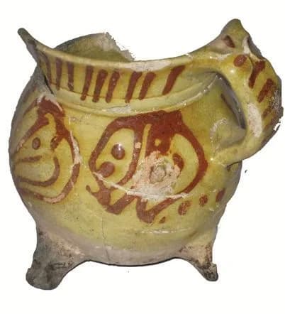 Dreibeiniger Henkeltopf als Scherbenfund, mit rostbraunem Malhorndekor auf gelber Glasur.