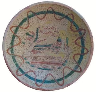 Schüssel mit liegendem Tier mit Federbusch im Spiegel, darunter ein Inschriftband, umgeben von grünem Ring und rotbraunem Wellenband.