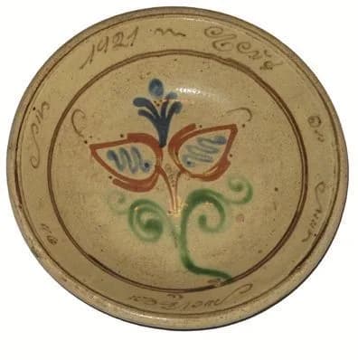 Teller mit stilisierter Blüte in Blau, Rot und Grün im Spiegel, am Rand eine umlaufende Inschrift mit der Jahreszahl 1921.