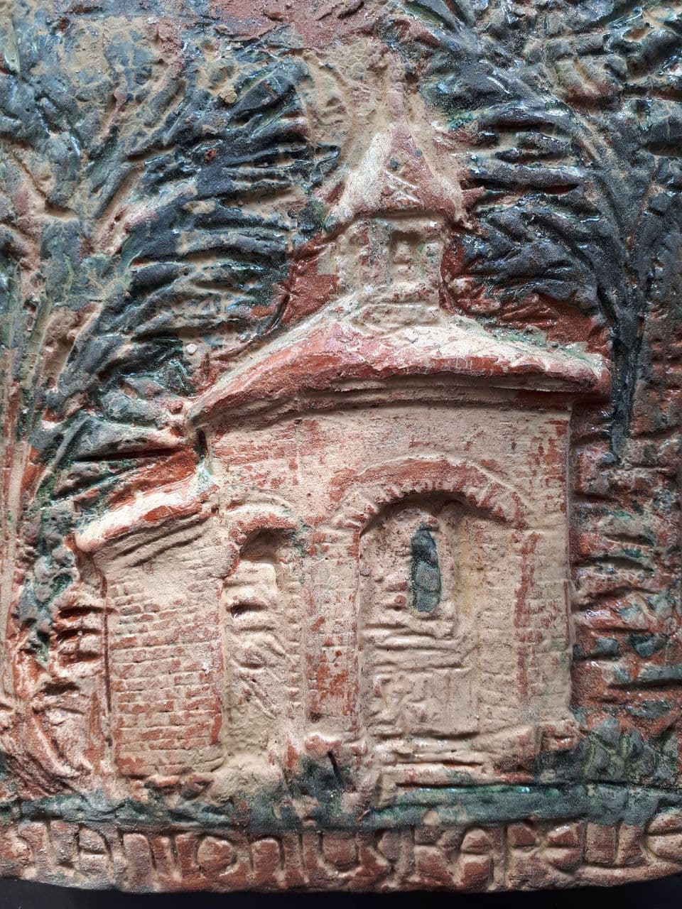 Detail eines Wandreliefs: Kapelle mit Dachreiter zwischen Bäumen, darunter der Schriftzug „Antoniuskapelle“, in Rotbraun, Beige und Graublau.