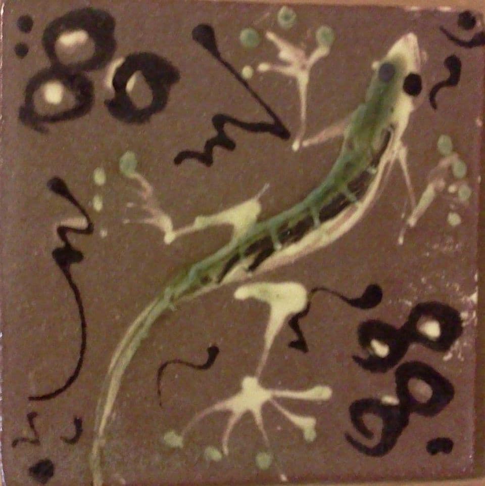Fliese mit grünem Gecko, hellen Spritzern und schwarzen Schnörkeln auf graubraunem Grund.
