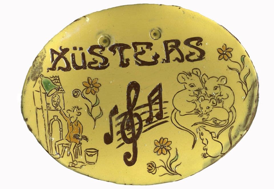 Ovales, gelb glasiertes Türschild mit dem Namen „Küsters“, Notenschlüssel, Mäusen, Blumen und einem Mann an einer Turmuhr mit Glocke.