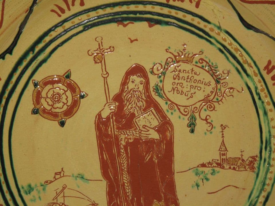 Teller mit dem heiligen Antonius mit Kreuzstab und Buch, daneben eine Rose und eine gekrönte Kartusche mit der Inschrift „Sancta Anthonius ora pro nobis“, im Hintergrund eine Kirche.