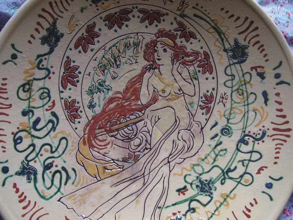 Detail eines Jugendstiltellers: sitzende Frau mit langem rotem Haar vor einem Kreis mit Vögeln, umgeben von dunkelroten Blüten und Ranken.