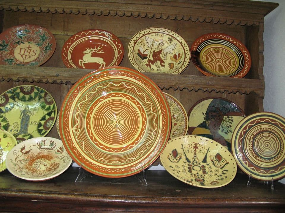 Holzregal mit dekorierten Tellern und Schüsseln, darunter Teller mit „IHS“, springendem Hirsch, geflügelten Figuren, Kreuz und Spiralmustern.