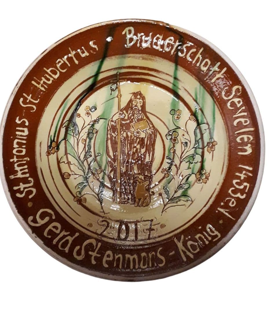 Königsteller der Bruderschaft Sevelen mit Heiligenfigur, Zweigen und Königsname in weißer Schrift auf braunem Rand, Jahr 2017.