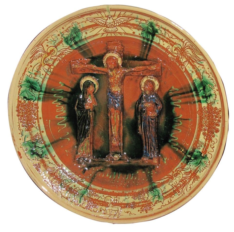Reliefteller mit Christus am Kreuz zwischen Maria und Johannes auf rotbraunem Grund mit grünen Glasurstrahlen, am Rand Taube, Weinlaub und Trauben.