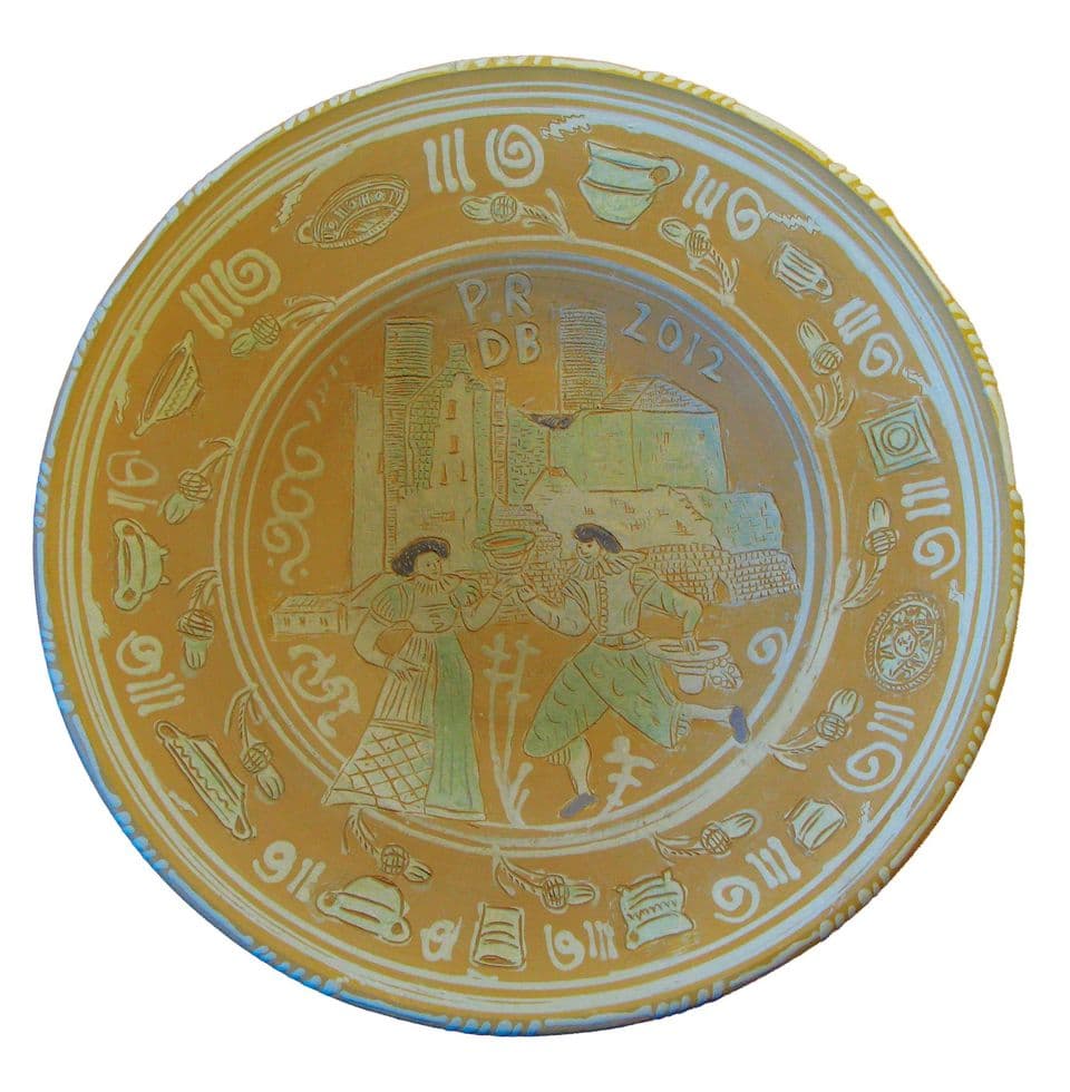 Ockergelber Teller mit einem Paar in historischer Kleidung vor einer Burg mit Türmen, darüber „P.R DB 2012“, am Rand Gefäße und Spiralen in heller Ritzzeichnung.